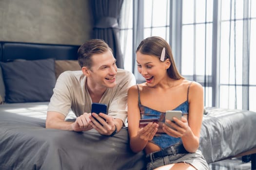 Young couple sit in the home bedroom using online payment app and digital wallet on smartphone to pay with credit card. E commerce shopping and modern purchasing via mobile internet. Unveiling