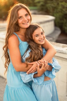 Portrait of mother and daughter in blue dresses with flowing long hair against the backdrop of sunset. The woman hugs and presses the girl to her. They are looking at the camera