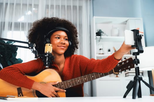 Host channel of beautiful African woman influencer setting smartphone, sing with play guitar in broadcast. Time slot of music blogger on live social media online. Concept of audio creator. Tastemaker.