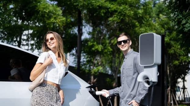 Young couple travel with EV electric car charging in green sustainable city outdoor garden in summer shows urban sustainability lifestyle by green clean rechargeable energy of electric vehicle innards