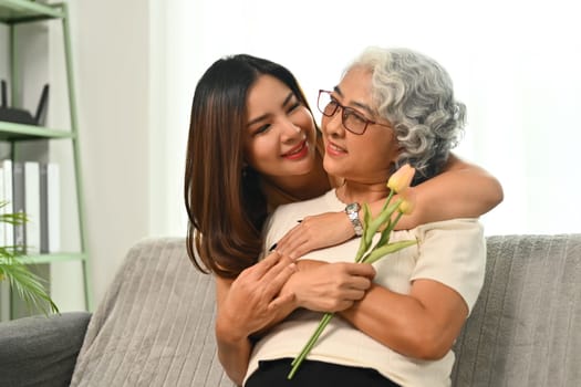 Smiling adult daughter giving beautiful spring tulips to her senior mother at home.