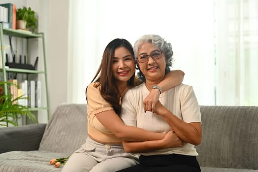 Smiling grey haired senior mother and young daughter sitting on couch at home, looking at camera.