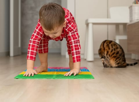 Sport and health concept. A little boy 4 years old in red checkered pajamas is having fun on a multi-colored massage orthopedic mat with spikes in a home interior. Close-up. Soft focus.