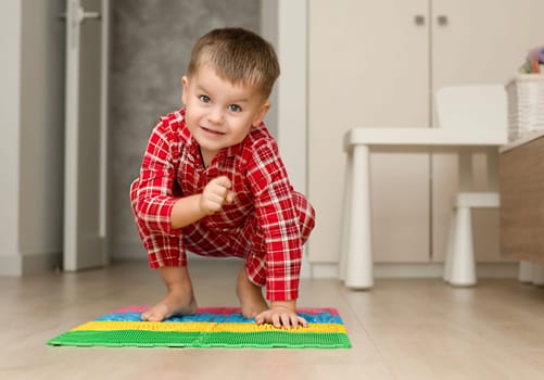 Sport and health concept. A little boy 4 years old in red checkered pajamas is having fun on a multi-colored massage orthopedic mat with spikes in a home interior. Close-up. Soft focus.