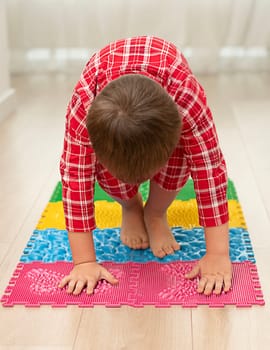 Sport and health concept. A little boy 4 years old in red checkered pajamas is having fun on a multi-colored massage orthopedic mat with spikes in a home interior. Close-up. Soft focus.