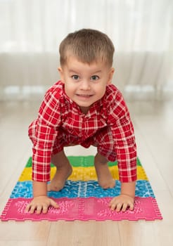 Sport and health concept. A little boy 4 years old in red checkered pajamas is having fun on a multi-colored massage orthopedic mat with spikes in a home interior. Close-up. Soft focus.