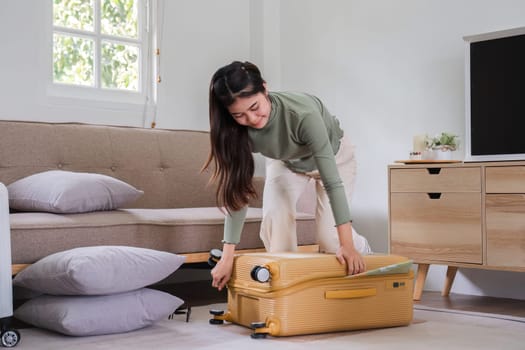 Beautiful Asian woman try to pack clothes into a yellow suitcase to prepare for a weekend getaway..