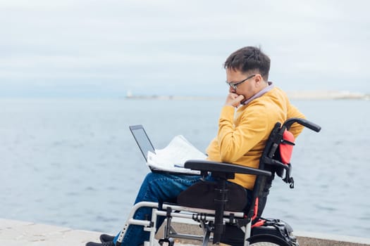 A man with disabilities sits with a laptop and papers, documents by the sea