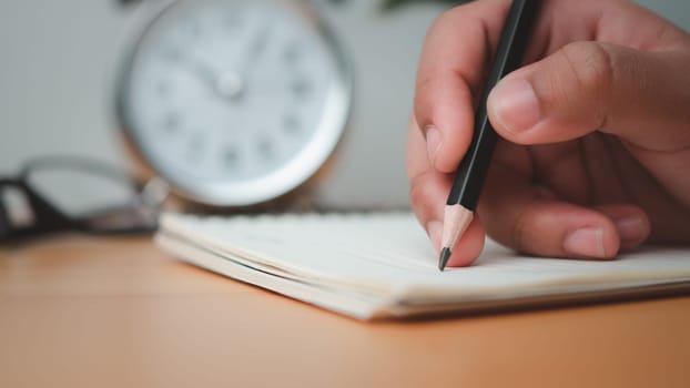 Human hands writing notes with pencil on paper on wooden table surface.