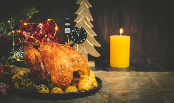 Hot chicken with potatoes in a dish, paper clock midnight, a bottle of champagne, a lit candle and a spruce branch with a garland on a black wooden table with copy space, close-up side view.