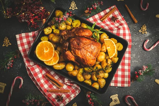 Chicken with potatoes, herbs, orange and cinnamon sticks in a baking dish on a kitchen red checkered napkin and christmas decoration on a dark background, close-up top view.
