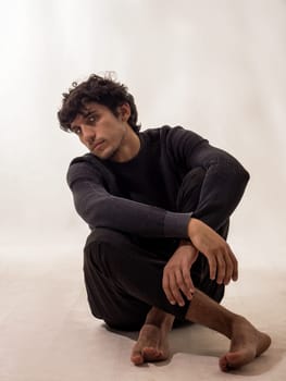 Serene barefoot handsome young man sitting on the ground with his legs crossed, in studio shot on white background.