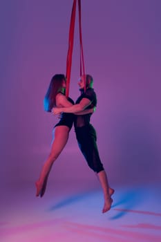 Gymnasts performance on a red canvases. Beautiful woman and strong man in a dark sport suits are looking at each other and doing acrobatic elements in a studio against a colorful background. Dancing in the air with balance.