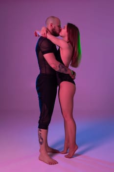 Pretty maiden and an athletic man in a dark sport suits are kissing each other while posing in a studio against a colorful background. Gymnasts performance.