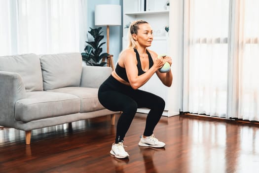 Athletic and sporty senior woman engaging in leg day training session with squat and bodyweight kettle ball at home exercise as concept of healthy fit body lifestyle after retirement. Clout