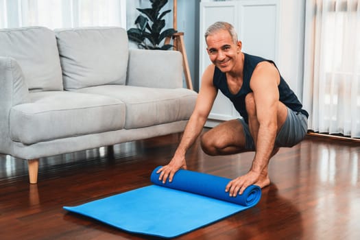 Active and sporty senior man preparing, rolling fitness exercising mat on living room floor at home. Home exercise as concept of healthy fit body lifestyle after retirement. Clout