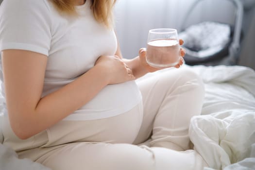 Pregnant woman sits on the bed, tenderly embracing her growing belly and holding glass of water.