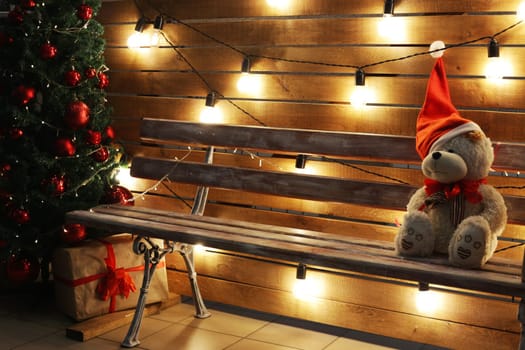 A sad teddy bear in a Santa hat sits on a bench next to a New Year tree, a gift box, against the backdrop of festive glowing lamps.