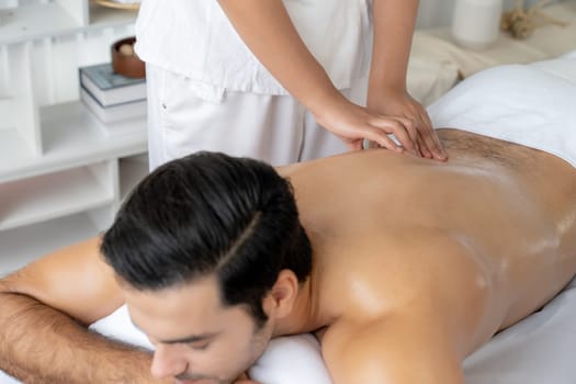 Caucasian man customer enjoying relaxing anti-stress spa massage and pampering with beauty skin recreation leisure in day light ambient salon spa at luxury resort or hotel. Quiescent