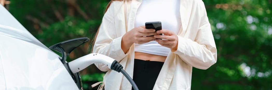 Young woman use smartphone to pay for electricity at public EV car charging station green city park. Modern environmental and sustainable urban lifestyle with EV vehicle. Panorama Expedient