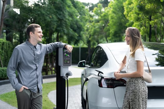 Young couple travel with EV electric car charging in green sustainable city outdoor garden in summer shows urban sustainability lifestyle by green clean rechargeable energy of electric vehicle innards