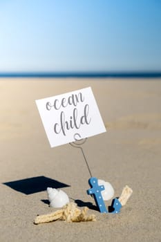 OCEAN CHILD text on paper greeting card in anchor paper holder and starfish seashell summer vacation decor. Sandy beach sun coast. Holiday concept postcard. Getting away Travel Business concept