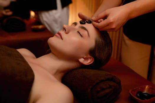 Caucasian couple enjoying relaxing anti-stress head massage with hot stone and pampering facial beauty skin recreation leisure in warm candle light ambient salon spa in resort or hotel. Quiescent