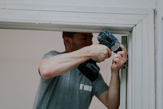 One handsome young male builder stands in a doorway and unscrews the screws of the sling with a drill, close-up side view. Concept of construction work.