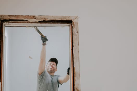 A young caucasian man in a uniform and gray textile gloves with real emotions on his face dismantles a board from a doorway with a crowbar and with rubbish spilling onto the floor, close-up view from below with selective focus. Construction work concept.