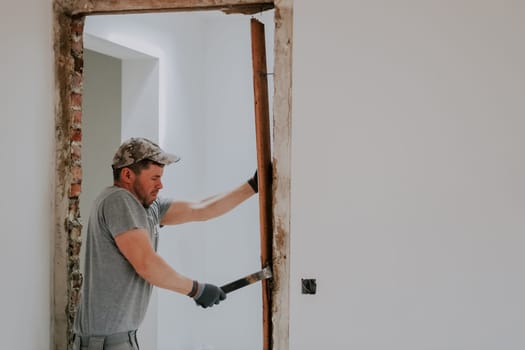 A young Caucasian man in a uniform and gray textile gloves emotionally dismantles planks from a doorway using a crowbar, close-up side view with selective focus. Construction work concept.