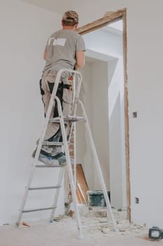 One young Caucasian man in a uniform and cap clears a doorway from old putty using a crowbar while standing from his back on a stepladder in a room with white walls, close-up side view with selective focus. Construction work concept.