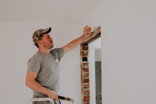 One young Caucasian man in uniform and cap clears the top beam of a doorway of old putty using one hand while standing sideways on a stepladder in a room with white walls, close-up side view with selective focus. Construction work concept.
