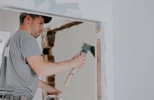 One young Caucasian man in uniform and cap clears old putty from the beams of a doorway with one hand using an ax, close-up side view with selective focus. Construction work concept.