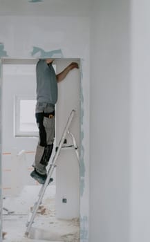 One young Caucasian man in a gray T-shirt clears old putty from the top of a doorway with one hand using an ax while standing sideways on a stepladder, close-up side view with selective focus. Construction work concept.