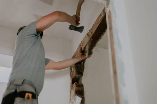 One young Caucasian man in a gray T-shirt and cap uses an ax to clear old putty from the top of a doorway with debris falling down, standing on a stepladder, close-up view from below with selective focus. Construction work concept.