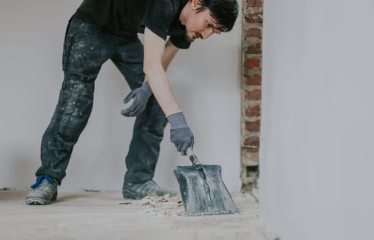 One young handsome brunette Caucasian woman in a black uniform stands, bending over, collecting construction debris from the floor with a shovel, close-up side view.