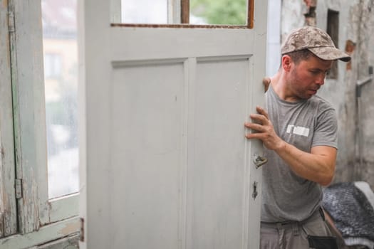 One young handsome Caucasian man in a gray T-shirt and cap holds with both hands an old wooden door while standing in a room where renovations are underway, close-up side view.
