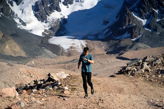 A young man runs along a trail in the snow-capped mountains, doing outdoor workout. The runner warms up, prepares for the race, leads a healthy active lifestyle.