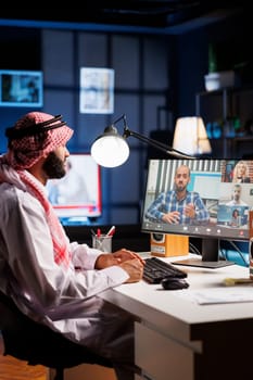 At the office table, Arab man dressed in traditional attire participates in online video meetings. Muslim guy pays close attention to colleagues while utilizing desktop pc for communication and study.