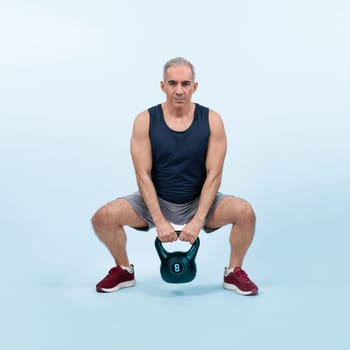 Full body length shot athletic and sporty senior man doing squat with kettlebell for body workout on isolated background. Healthy active physique and body care lifestyle after retirement. Clout