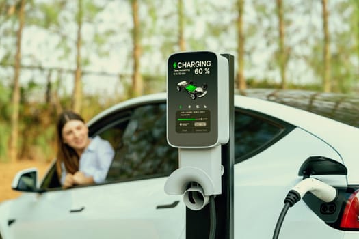 Focused EV charging station for electric vehicle's battery recharging on blurred background of young woman during her autumnal road trip travel. Eco friendly vacation with EV car during autumn. Exalt