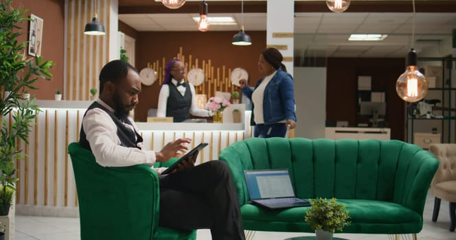 African american staff making online bookings on tablet in lobby, working at luxurious hotel and providing excellent concierge service for guests upon their arrival. Bellboy in uniform manage rooms.