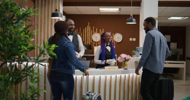 Wife and husband enter resort lobby, talking about front desk staff about room service and ammnities. Couple arriving in lounge area reception with suitcases, honeymoon vacation.