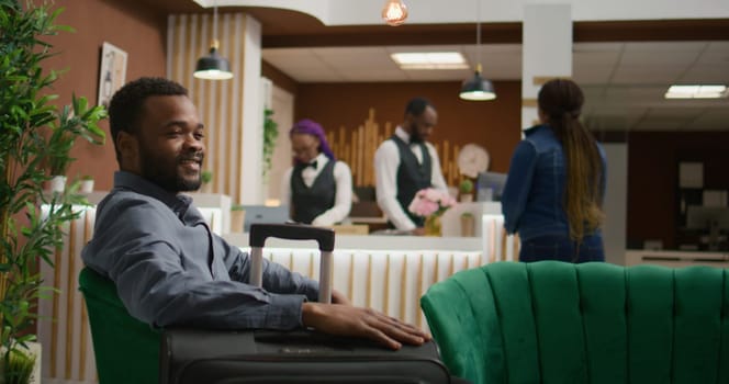 African american guest sit in lobby, preparing to do check in and enjoy hotel services, relaxing at reception after long flight. Person relaxes in lounge area at holiday retreat, tourism.