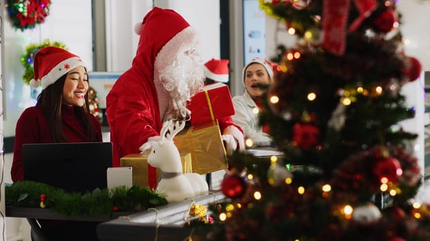 Multiethnic workers receiving gifts from colleague pretending to be Santa in xmas decorated office. Employee wearing costume surprising company personnel with presents during winter holiday season