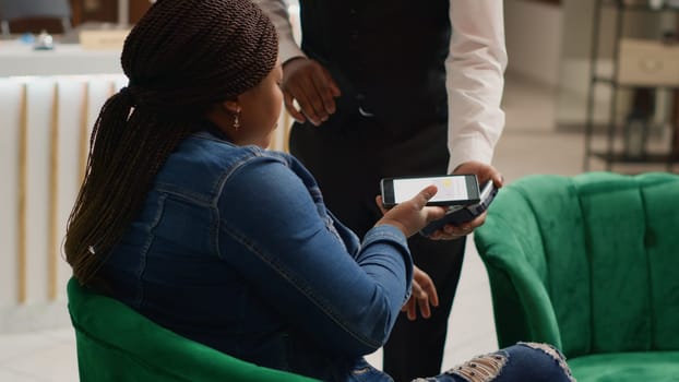 Client makes nfc payment at pos to pay for coffee cup served while waiting on room vacancy in hotel lobby. African american woman talking to bellhop about luxurious service provided. Handheld shot.