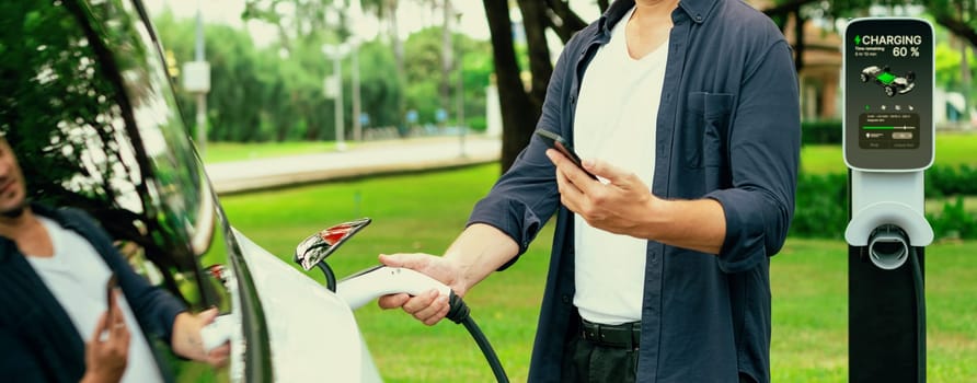 Man using smartphone online banking application to pay for electric car battery charging from EV charging station during vacation road trip at national park or summer forest. Panorama Exalt