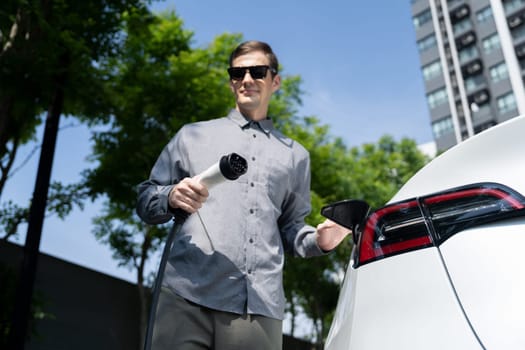 Young man travel with EV electric car charging in green sustainable city outdoor garden in summer shows urban sustainability lifestyle by green clean rechargeable energy of electric vehicle innards