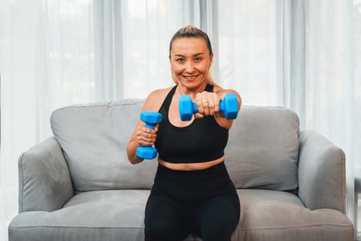 Athletic and sporty senior woman engaging in body workout routine, sitting on sofa and lifting dumbbell at home as concept of healthy fit body with body weight lifestyle after retirement. Clout