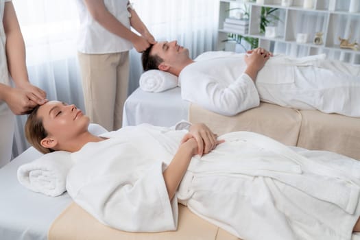 Caucasian couple enjoying relaxing anti-stress head massage and pampering facial beauty skin recreation leisure in dayspa modern light ambient at luxury resort or hotel spa salon. Quiescent